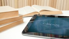 Das Foto zeigt ein Tablett, auf dem das Wort "e-learning" abgebildet ist. Das Tablett liegt gemeinsam mit einigen Büchern auf einem Tisch.