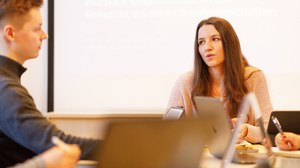 Man sieht eine junge Frau, die einem jungen Mann etwas erklärt. Vor ihnen stehen Laptops.