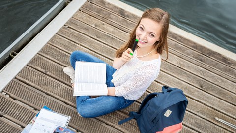 Lächelnde Studentin lernt auf einem Bootssteg an der Elbe. Sie blickt in die Kamera.