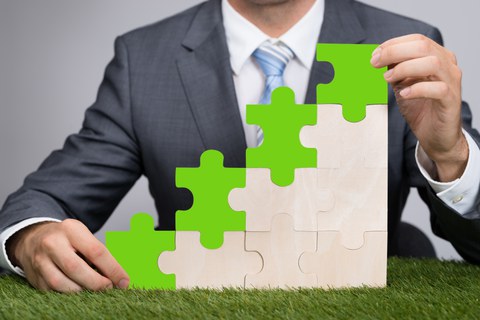 Das Foto zeigt eine Person mit Anzug, welche mit Puzzleteilen eine Art Treppe aufgebaut hat. Die obersten Teile sind grün. Der Untergrund, auf dem die Treppe steht, ist eine Wiese.