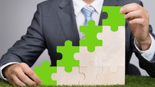 Das Foto zeigt eine Person mit Anzug, welche mit Puzzleteilen eine Art Treppe aufgebaut hat. Die obersten Teile sind grün. Der Untergrund, auf dem die Treppe steht, ist eine Wiese.