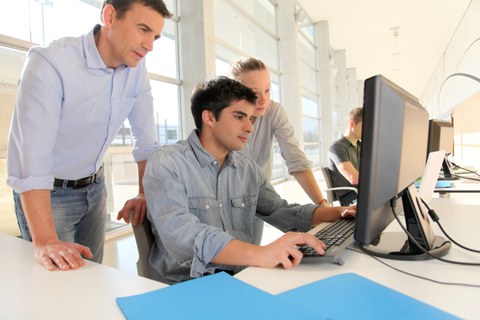 Das Foto zeigt zwei Studierende vor einem Computer. Der Junge sitzt auf dem Stuhl und tippt auf der Tastatur. Der Dozent steht daneben und schaut ebenfalls auf den Bildschirm. 