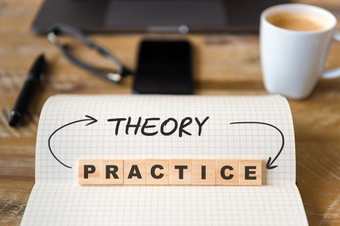 Das Foto zeigt einen karierten Block mit dem geschriebenen Wort "theroy". Mit aufgestellten Holzsteinen ist außerdem das Wort "practice" dargestellt. Die beiden Worte sind durch Pfeile miteinander verbunden. Im Hintergrund erkennt man verschwommen einen Büroschreibtisch.