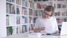 Das Foto zeigt eine Person am Schreibtisch. Sie liest in einem Buch und hat einen Stift zwischen die Zähne geklemmt. Hinter ihr steht ein Bücherregal.