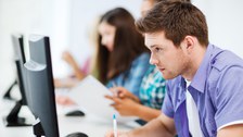 Das Foto zeigt mehrer Personen, die vor ihren Computern sitzen. Im Vordergrund erkennt man einen jungen Mann, der gerade sehr angestrengt auf seinen Bildschirm schaut. 