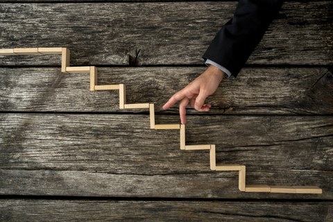 Das Foto zeigt einen Arm vor einem hölzernen Hintergrund. Darauf liegen Holzstückchen, die eine Treppe bilden. Die Person geht mit ihren Fingern die Treppe hinauf. 
