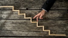 Das Foto zeigt einen Arm vor einem hölzernen Hintergrund. Darauf liegen Holzstückchen, die eine Treppe bilden. Die Person geht mit ihren Fingern die Treppe hinauf. 