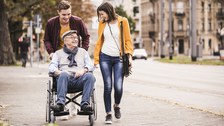 Foto zeigt einen Mann und eine Frau, die mit ihrem, im Rollstuhl sitzenden, Großvater spazieren gehen