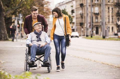 Foto zeigt einen Mann und eine Frau, die mit ihrem, im Rollstuhl sitzenden, Großvater spazieren gehen