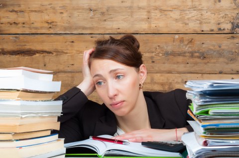Das Foto zeigt eine Frau mit frustriertem Gesichtsausdruck. Sie sitzt zwischen zwei großen Bücher- und Hefterstapeln und hat weitere Dokumente vor sich liegen.