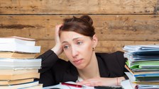 Das Foto zeigt eine Frau mit frustriertem Gesichtsausdruck. Sie sitzt zwischen zwei großen Bücher- und Hefterstapeln und hat weitere Dokumente vor sich liegen.