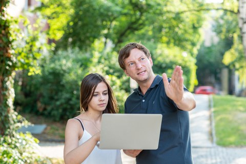 Foto von zwei Personen im Freien, die ein Laptop halten. Der Mann zeigt auf etwas vor ihnen.