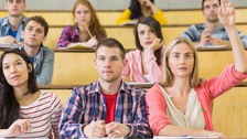 Das Foto zeigt mehrere Studierende während einer Veranstaltung in einem Hörsaal. Eine Studentin hebt gerade ihre Hand.