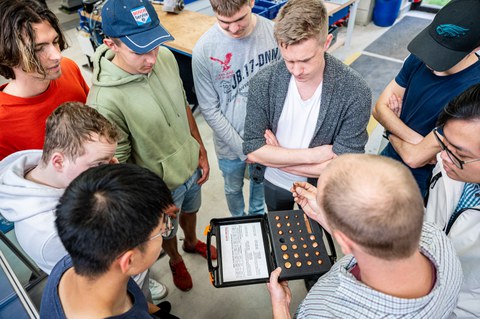 Foto einer Gruppe Studierender und ihrem Dozenten aus der Vogelperspektive. Der Dozent erklärt seinen Studenten was Schweißköpfe sind und hält dabei einen in der Hand.