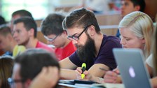 Das Foto zeigt einen Studenten inmitten eines vollen Hörsaals. Er schaut auf seinen vor sich liegenden Hefter.