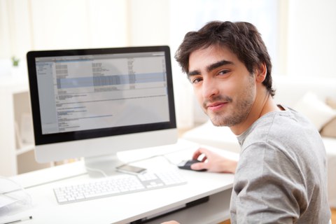 Foto: Ein junger Mann arbeitet an seinem Computer. Er lächelt und sieht glücklich aus.