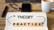  Das Foto zeigt einen karierten Block mit dem geschriebenen Wort "theroy". Mit aufgestellten Holzsteinen ist außerdem das Wort "practice" dargestellt. Die beiden Worte sind durch Pfeile miteinander verbunden. Im Hintergrund erkennt man verschwommen einen 