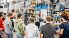 Foto einer Gruppe Studierender und ihrem Dozenten. Die Studenten beobachten im Halbkreis eine Flipchartpräsentation ihres Dozenten.