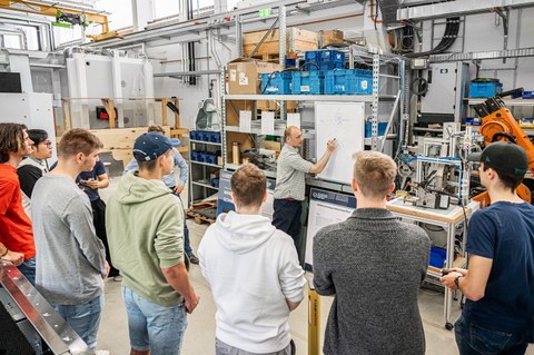 Foto einer Gruppe Studierender und ihrem Dozenten. Die Studenten beobachten im Halbkreis eine Flipchartpräsentation ihres Dozenten.