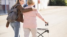 Foto zeigt eine ältere Frau, die mit ihrer Tochter an der Seite mit einem Rollator spazieren geht.
