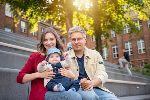 Foto eines studentischen Paares, das mit einem etwa 1-jährigem Kind auf dem Schoß auf den Fritz--Förster-Bau-Treppen sitzt.