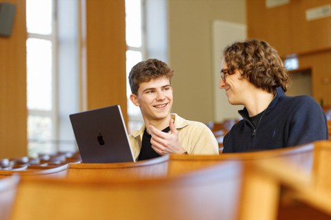 Man sieht zwei Studenten im Hörsaal, die sich unterhalten