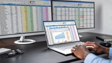 Foto einer Person, die an seinem Schreibtisch an 2 Bildschirmen und einem Laptop Tabellen und Diagramme bearbeitet