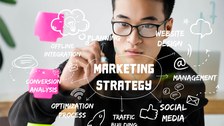Das Foto zeigt einen jungen Mann, der mit einem Stift an eine durchsichtige Tafel schreibt. Darüber liegt eine Mindmap, die sich um den Begriff "Marketing Strategy" dreht. Sie betrachtet unterschiedlkiche Aspekte, wie "planning", "social media" und "optimization process".