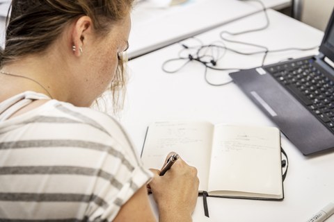 Foto einer Frau, die an einem Arbeitsplatz mit Laptop sitzt und etwas in ein Notizbuch schreibt