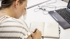 Foto einer Frau, die an einem Arbeitsplatz mit Laptop sitzt und etwas in ein Notizbuch schreibt
