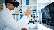 Foto eines Mannes im weißen Laborkittel, der vor einem Bildschirm sitzt und durch eine VR-Brille sieht