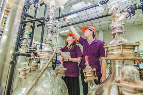 Zwei Studierende stehen in einem Labor. Sie tragen orangene Helme und violette Kittel. Im Vordergrund befinden sich zwei große Glasglocken, die über Rohre und Schläuche miteinander verbunden sind. 