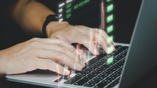 Foto zeigt die Hände eines Mannes auf einer Laptoptastatur mit eingeblendeten Graphiken und Zahlen. 