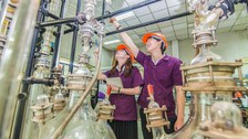 Zwei Studierende stehen in einem Labor. Sie tragen orangene Helme und violette Kittel. Im Vordergrund befinden sich zwei große Glasglocken, die über Rohre und Schläuche miteinander verbunden sind. 