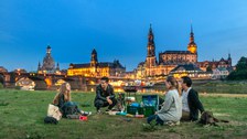 Foto: Vier Studenten sitzen mit einem Hund am Abend an den Elbwiesen von Dresden und grillen.