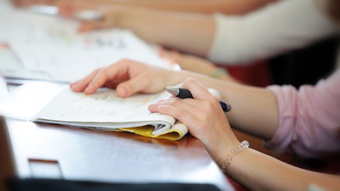 Foto von zwei Händen mit einem Stift in der linken Hand auf einem Hefter im Hörsaal