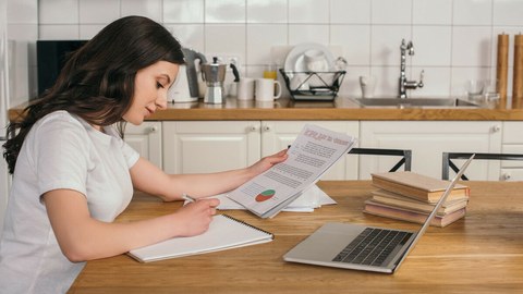 Das Foto zeigt eine SDas Bild zeigt eine Frau an einem Küchentisch. Vor ihr steht ihr Laptop, sie hält Zettel in der Hand und notiert etwas auf einem Block. 