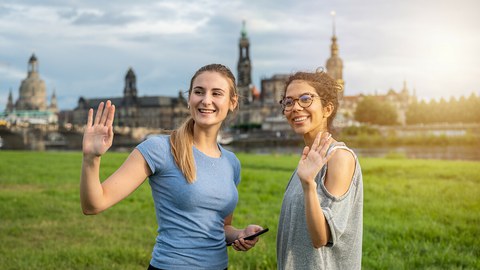 Foto zeigt zwei lächelnde und winkende Studentinnen an der Elbe.