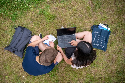 Bild von zwei Studenten, die auf der Wiese zusammen am Laptop arbeiten.