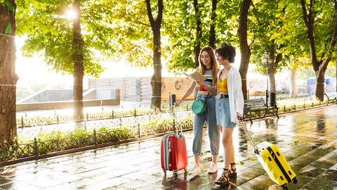 Foto von zwei fröhlichen schönen Frauen, die lächeln und eine Karte in der Hand halten, während sie mit Koffern am grünen Stadtboulevard spazieren