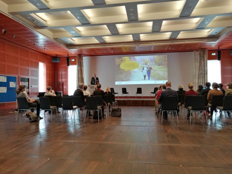Personen sitzen in einem Saal, vorn steht ein Redner