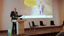 Ein Redner spricht vor einer Präsentation. Auf der Folie sind 2 ältere Menschen zu sehen und die Aufschrift "Willkommen zum Datensalon"