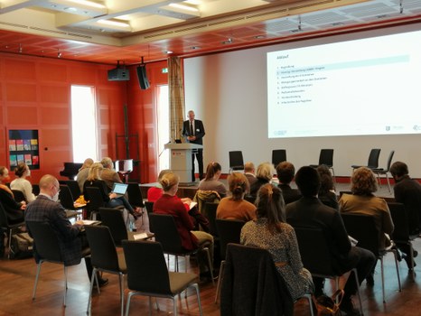 Personen sitzen in einem Saal, vorn steht ein Redner