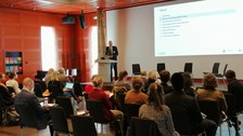 Personen sitzen in einem Saal, vorn steht ein Redner