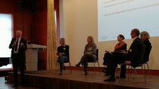 Fünf Personen sitzen auf einer Bühne, davor steht ein Redner