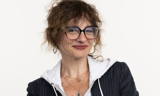 Woman with large black glasses, curly brown hair, red lipstick, black and white jacket, smiling at the camera