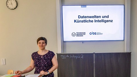 Eine weiße, weibliche Person mit kurzen Haaren steht vor einer Tafel und Folie mit dem Titel "Datenwelten und Künstliche Intelligenz"