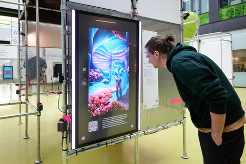 Eine Person betrachtet einen Bildschirm auf dem eine futuristisch aussehende Figur in einer Landschaft zusehen ist, die an den Meeresgrund erinnert. 