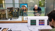 Zwei Personen betrachten einen Ausstellungsbereich. Eine Person im Vordergrund rechts hört einen Audiobeitrag der Ausstellung an.