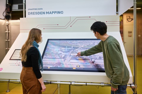 Zwei Personen stehen vor einem Bildschirm, der einen digitalen Stadtplan zeigt. Die Person rechts im Bild bedient den Bildschirm mit der Hand.